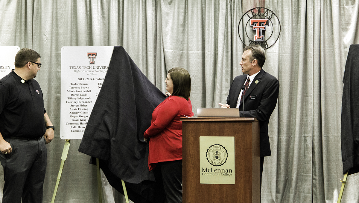 Lewis Snell stands behind a brown podium while looking over his right shoulder as a poster is unveiled from under a black cloth by a woman wearing a red sweater
