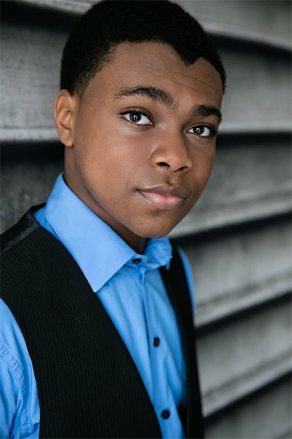 A headshot of Chase Greer leaning against a wall while wearing a blue buttoned down shirt with a black vest.
