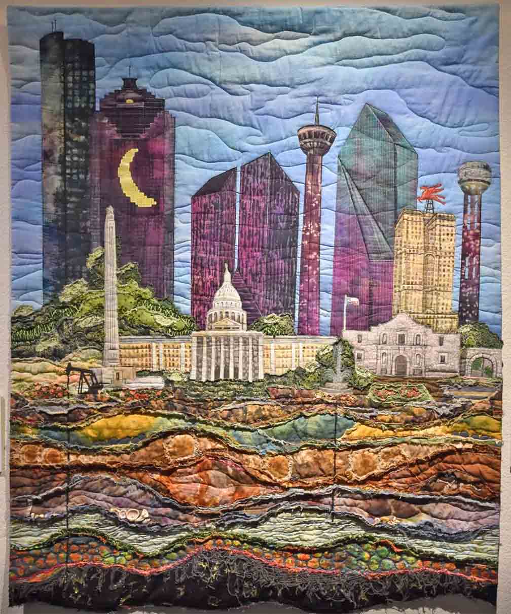vertically oriented art quilt with images of Texas buildings such as the Alamo and Capital sitting on the oil rich sub layers of dirt.