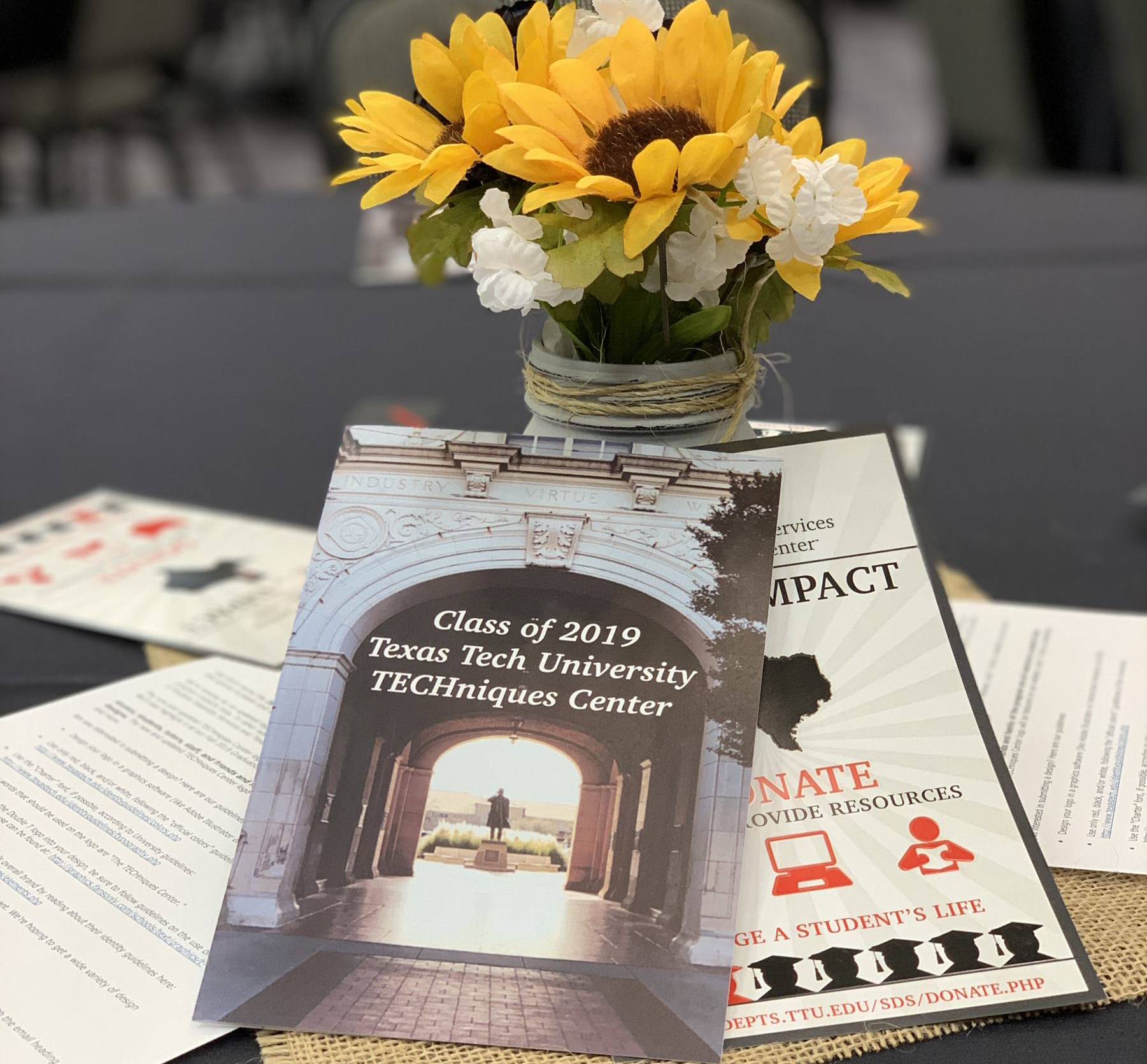 Pamphlets on a table focusing on the top one which says "Class of 2019 Texas Tech University Techniques Center" where a flower pot next to it with yellow and white flowers, all on a black table cloth. 