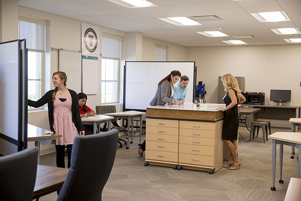 three people around desk, another writing on white board, a fifth working separately