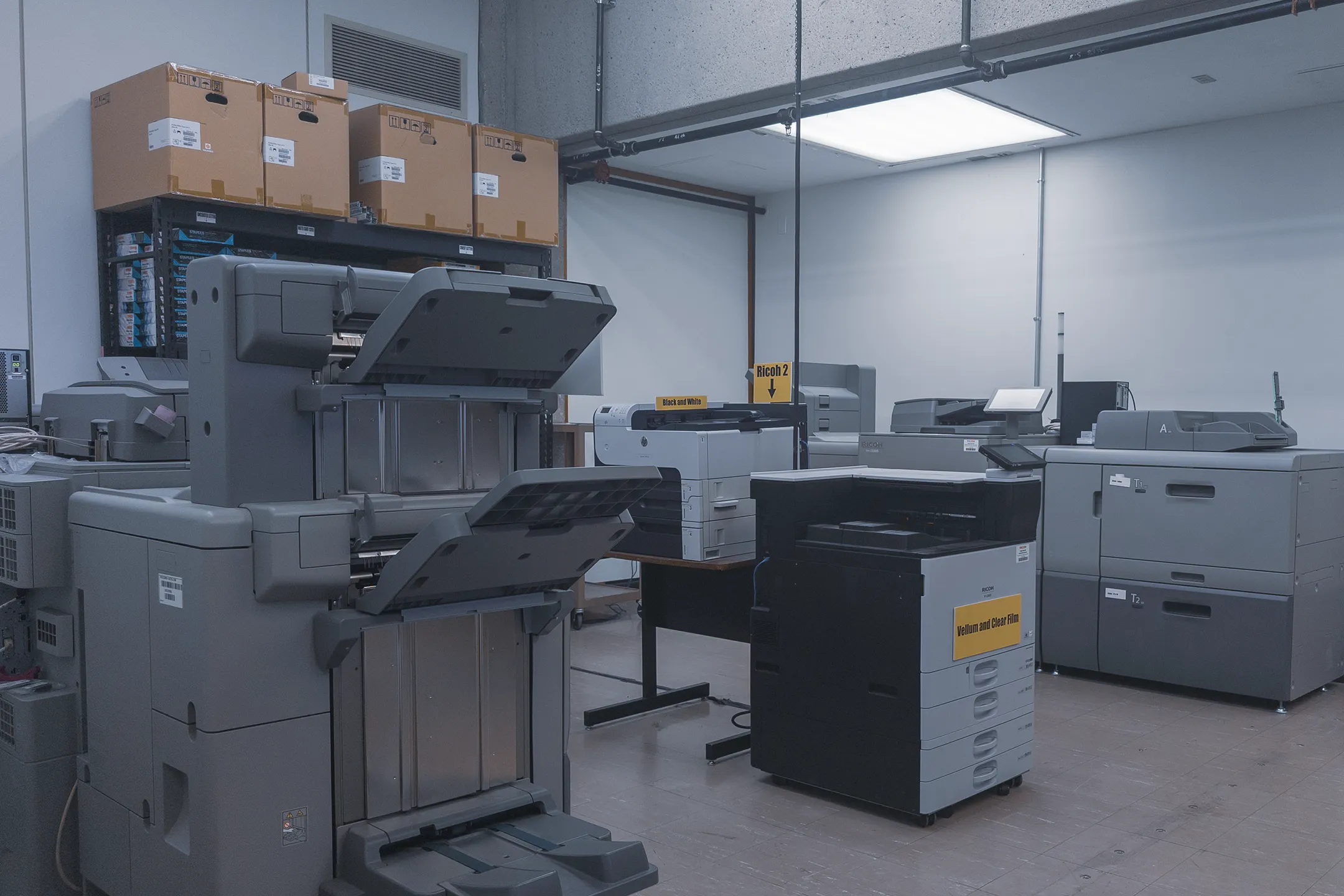 Photo of the print shop interior. Shelving full of paper against the back wall, with two large printers creating a U-shape with the space.