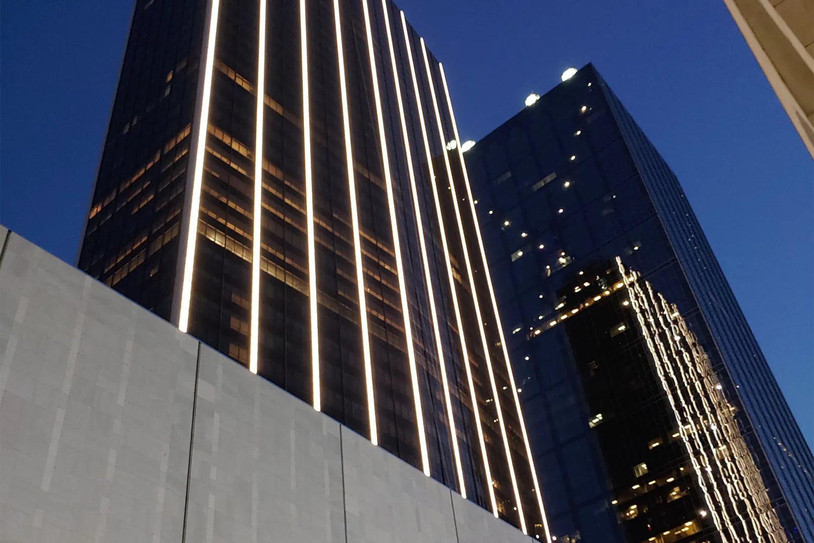 A city skyline at night. Photograph pointed upward at two large glass buildings, with the concrete awing of a third building in the bottom left.