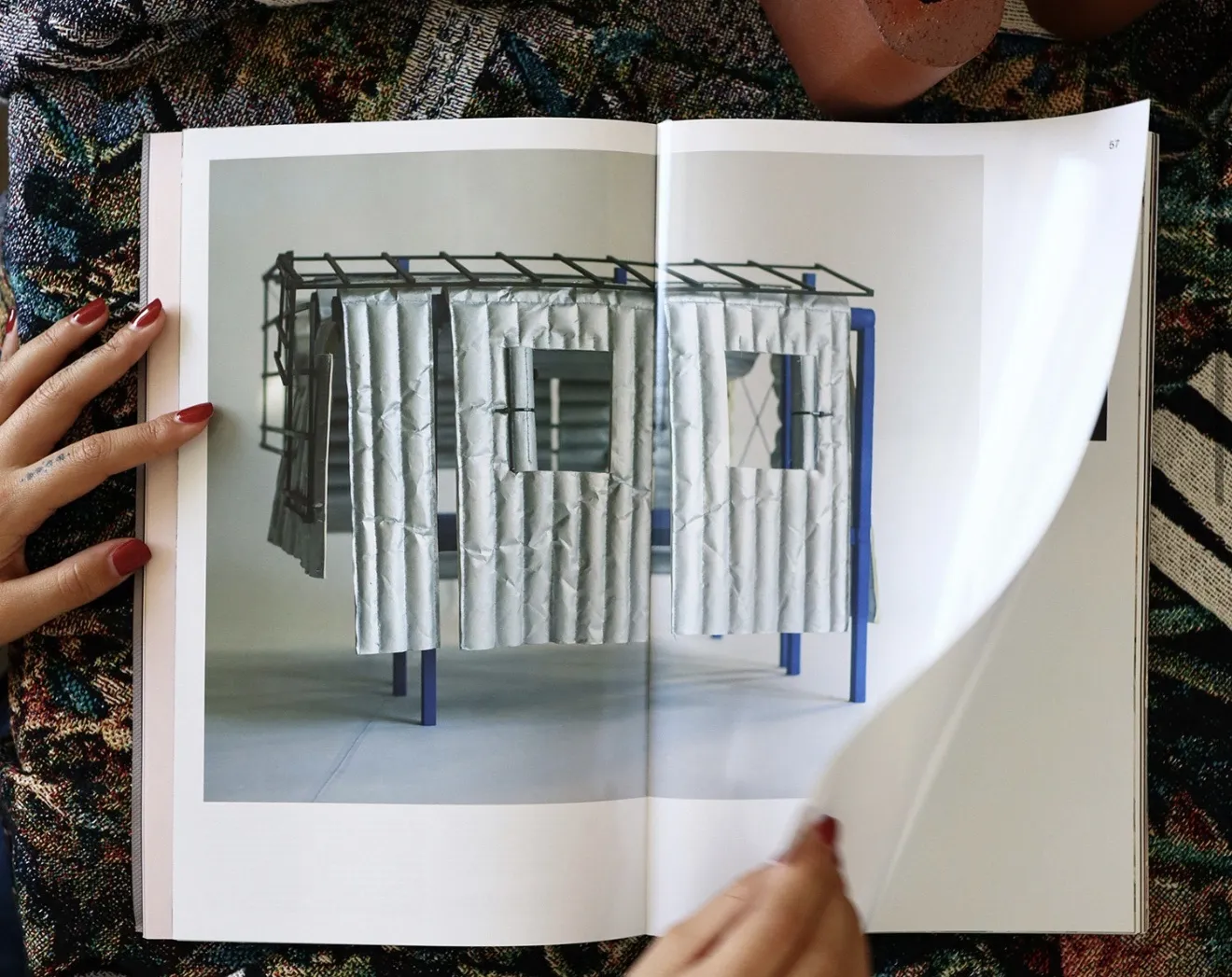 Hands holding a book and turning a page. The book is displaying an image of a basic home structure made from blue piping and metallic cloth.