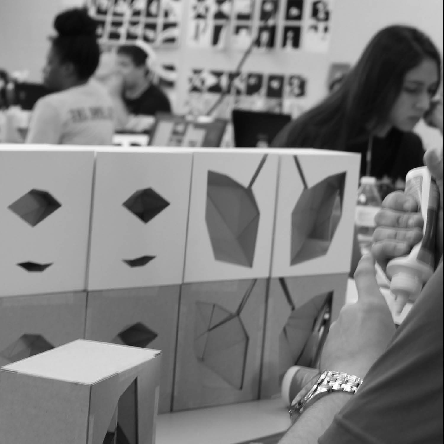 Student applying glue to a paper model, with other students working on projects in the background.