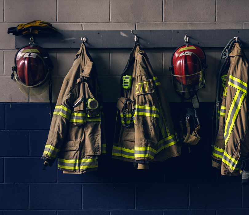 Row of fire service gear hanging on a wall, including several tan turnout coats with reflective stripes and red helmets.