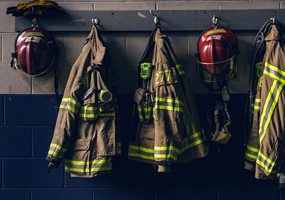 Row of fire service gear hanging on a wall, including several tan turnout coats with reflective stripes and red helmets.