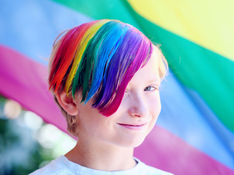 child with rainbow colored hair in front of a rainbow flag