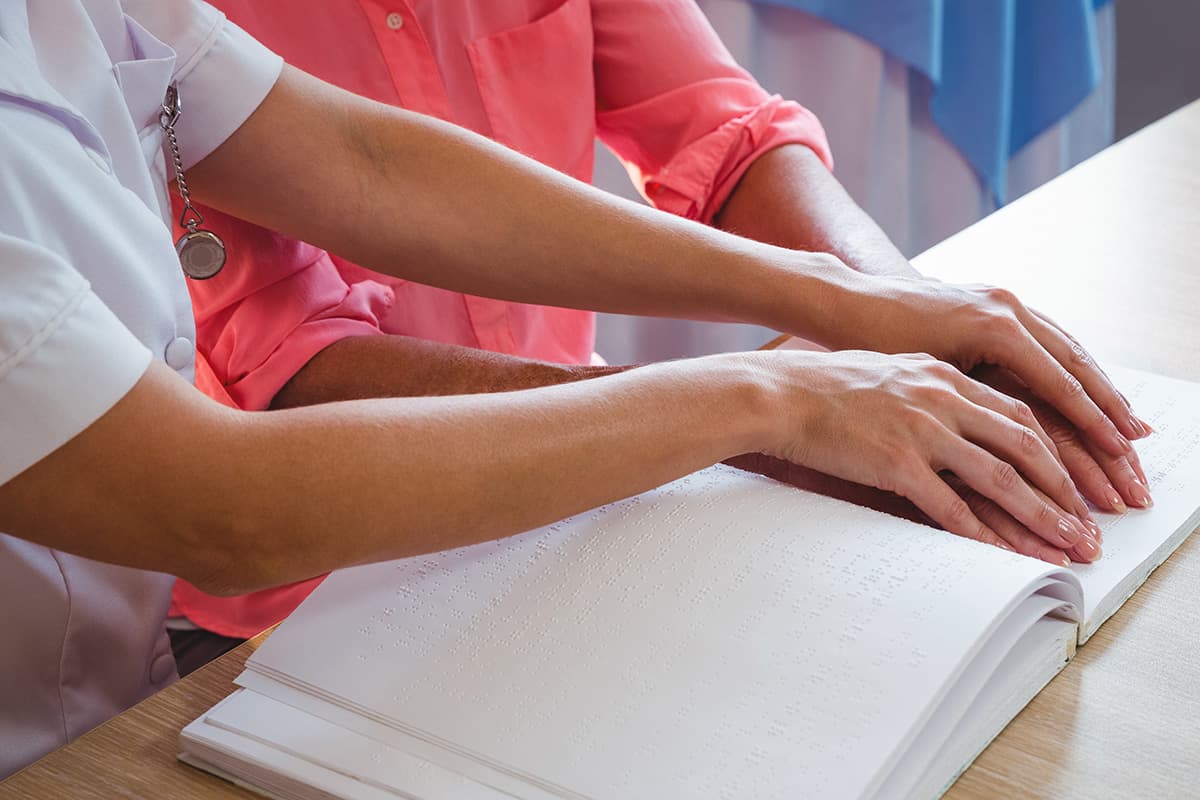 Woman reading from a Braille book with teacher's help.