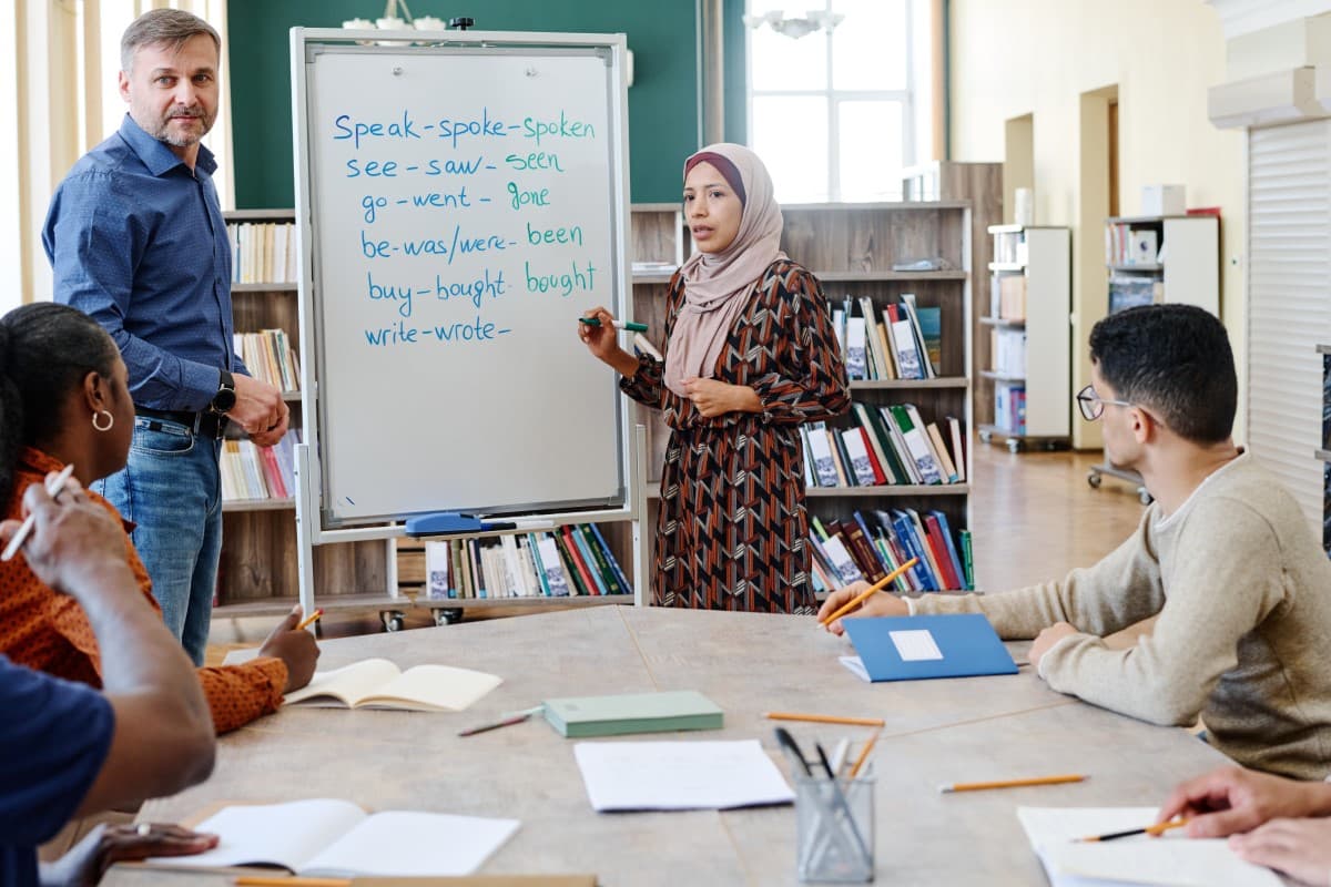 An english teacher and student diagram irregular english verbs at a whiteboard in a library classroom.
