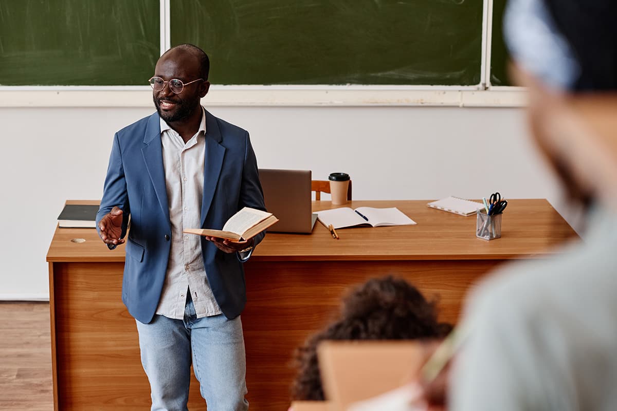 Lecturer presenting class material in front of desk and chalkboard.