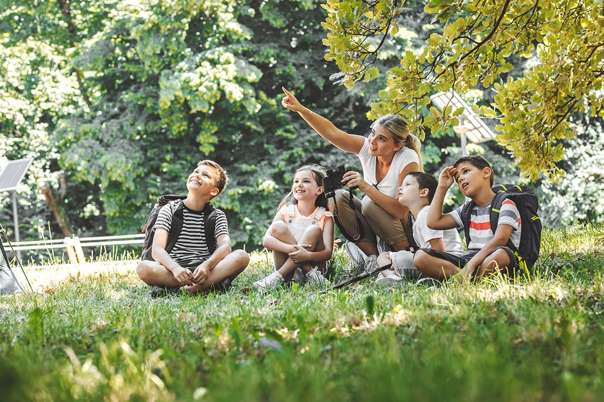 Teacher and young students having class outdoors under a tree.