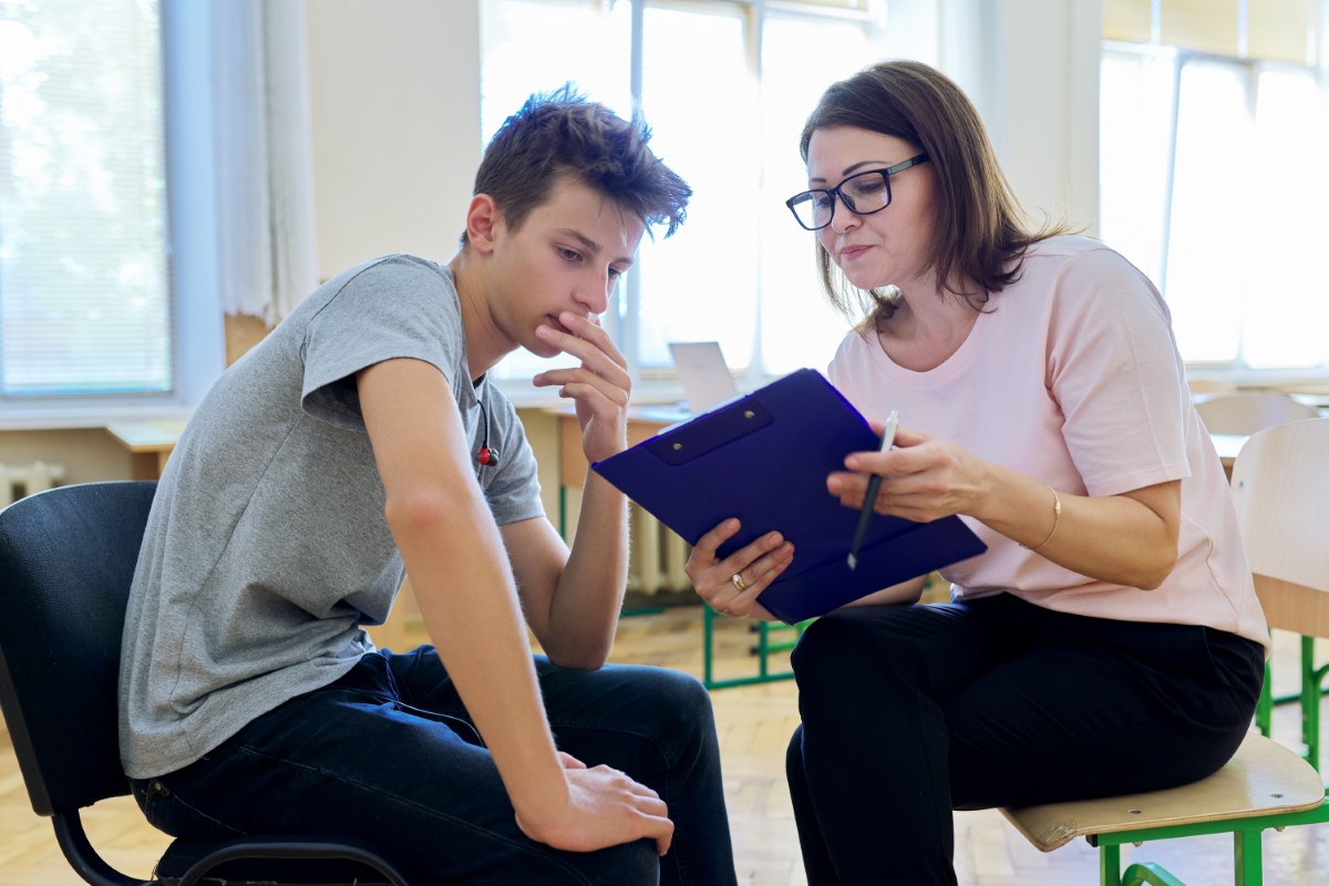 A school psychologist and male high school student examine documents on a clipboard together.