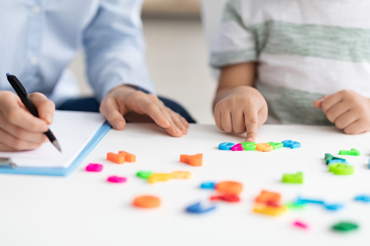 Teacher with pen and clipboard assisting a young student with letter and number shapes.