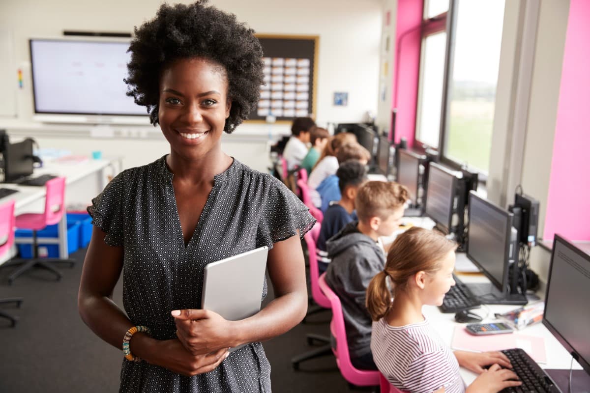 Teacher posing holding a tablet in front of a line of students at computers