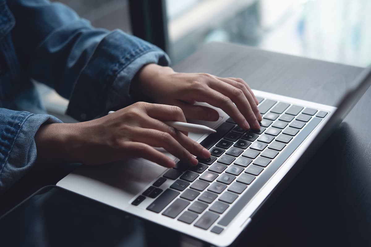 Closeup of hands typing on laptop while holding an Apple Pencil.