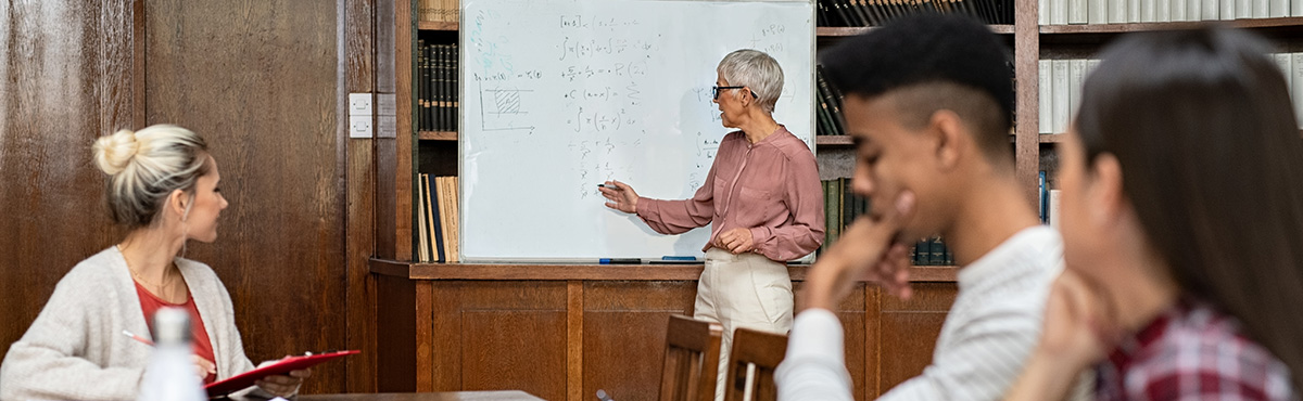 Banner image - photo in library of woman at a whiteboard instructing a group of three college-age students