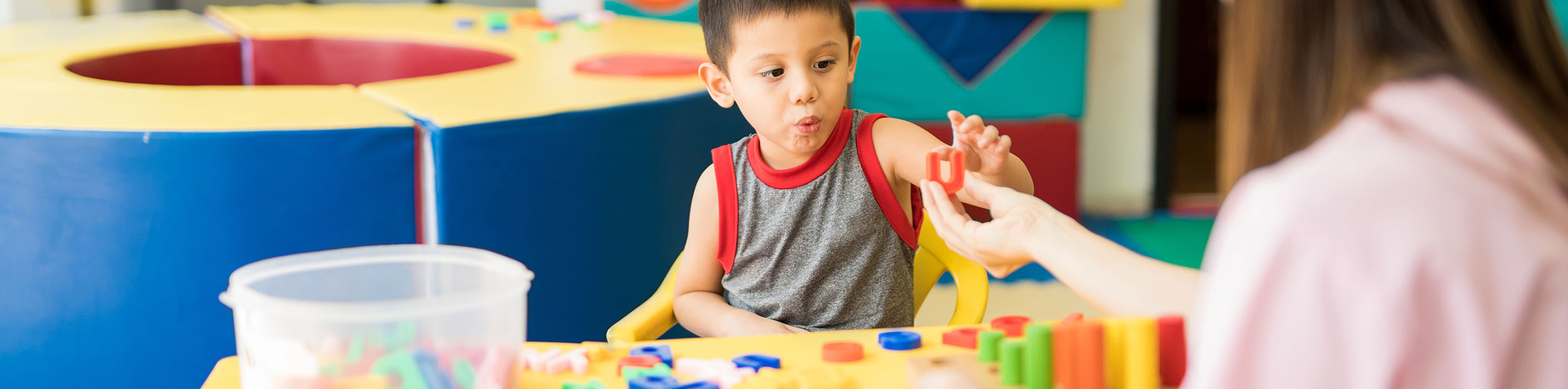 Photo of a child working with colorful plastic letters. A teacher hands him a letter.