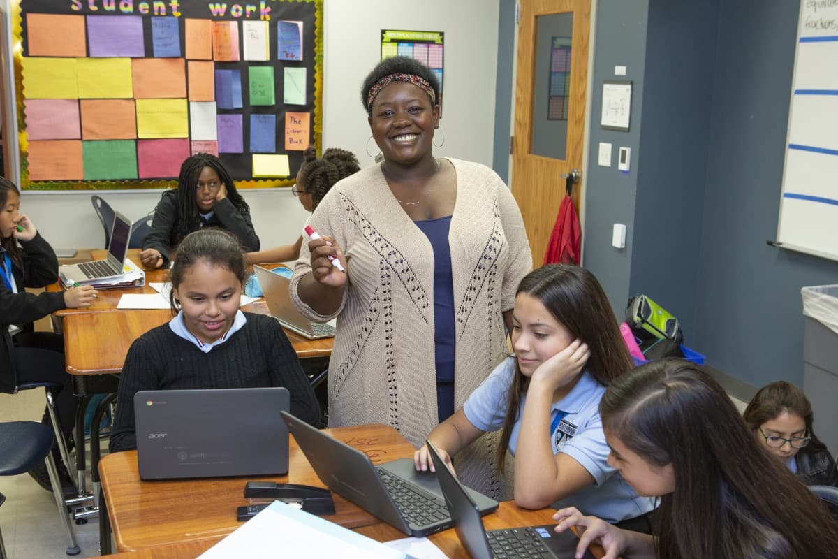 Teacher smiling at the camera while middle school students work on laptops in a classroom.