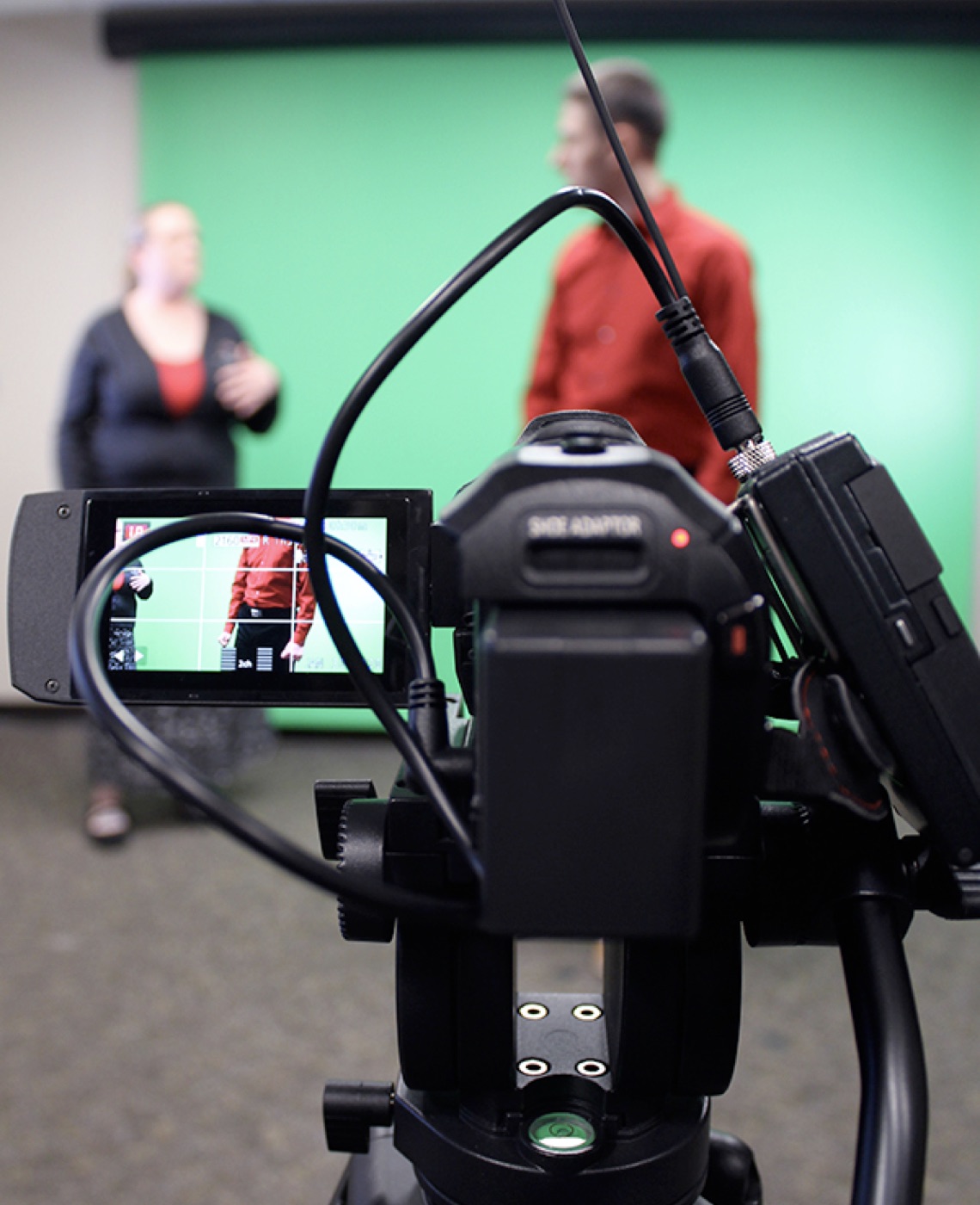 A man wearing a red shirt and a woman wearing a navy blue sweater stand in front of a green screen in the background of the photo while a black camera with a red light rests in the foreground.