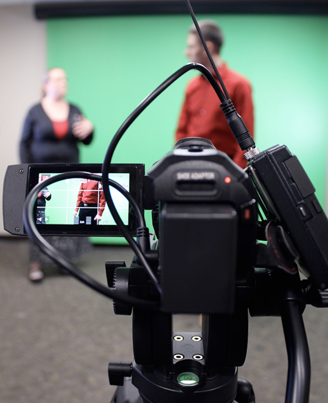 The large green screen allows for a variety of animation and enhancement options in film production. A man wearing a red shirt and a woman wearing a navy blue sweater stand in front of a green screen in the background of the photo while a black camera with a red light rests in the foreground.