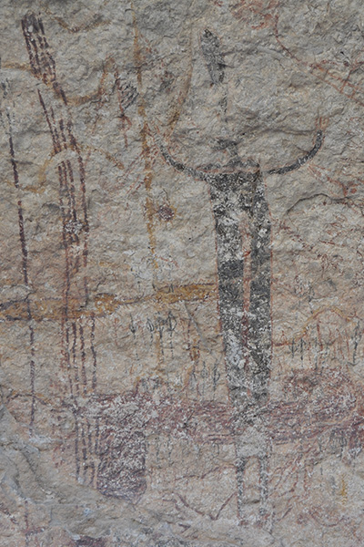 A cave drawing on brown rock that displays what appears to be a tall human towering over their surrounding environment.