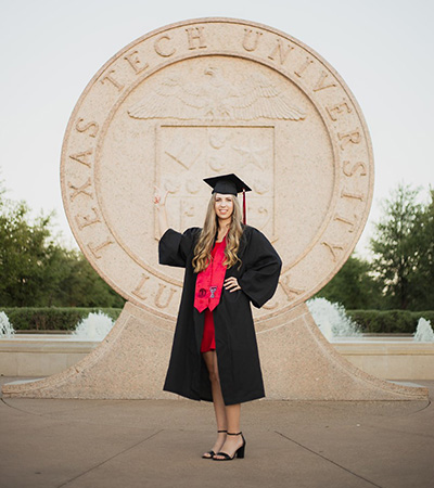 A woman stands in front a structure of the Texas Tech seal while wearing a Texas tech cap and gown and posing.