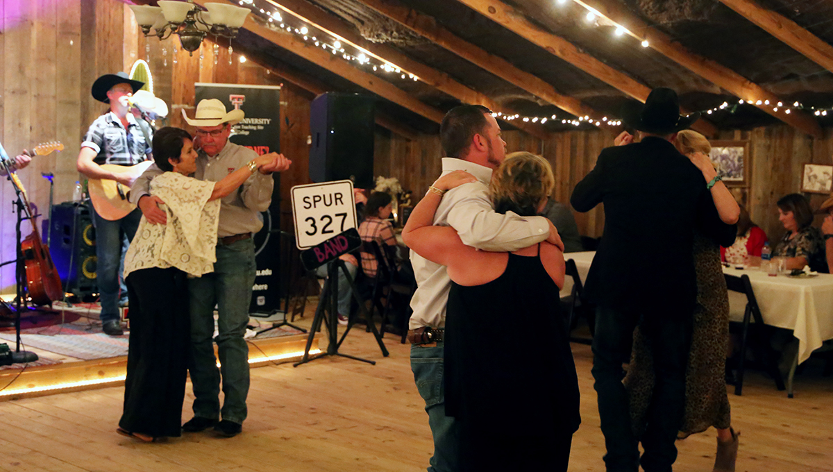 Couples dance together indoors on top of a wooden floor surrounded by bright lights and other sitting patrons