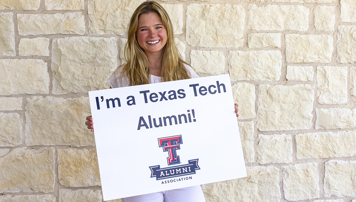 Madison stands in front of a light-colored brick building while holding up a sign that says I'm a Texas Tech Alumni