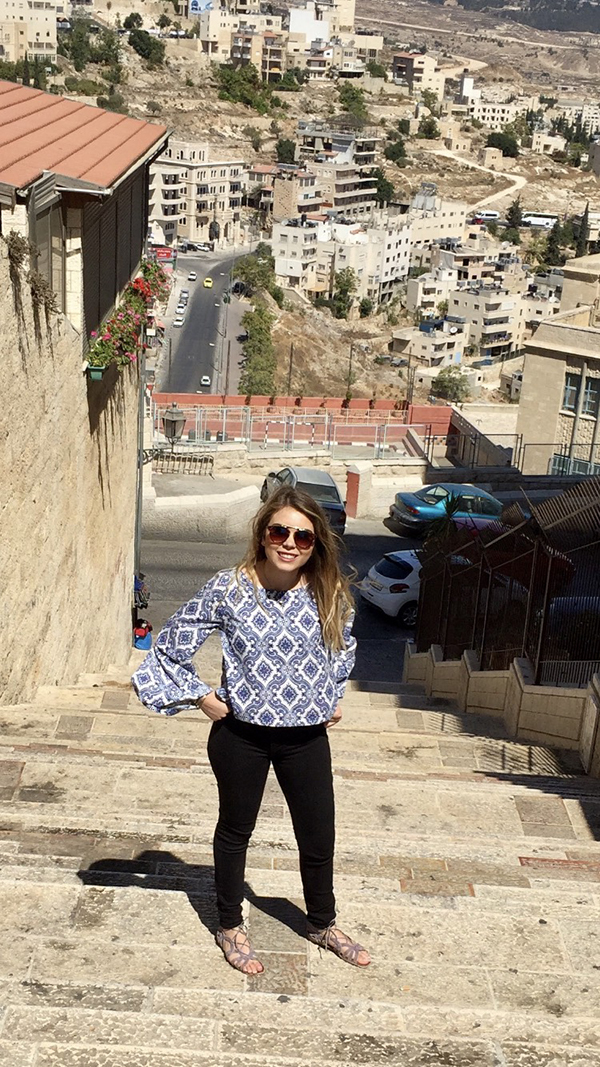 Daniella Munoz stands on the top step of an outside staircase while smiling with scenic Spain in the background