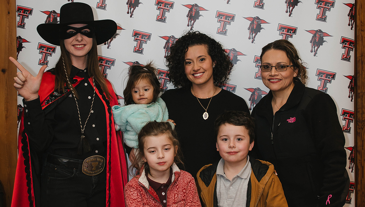 Tabitha holds her youngest daughter in her right arm while standing behind her son and daughter and in between her mother and the masked rider 