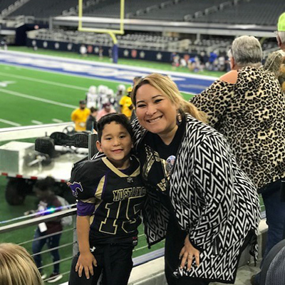 A woman with blond hair smiles and leans against her young son who wear a football uniform and pads as both pose with a football field in the background.