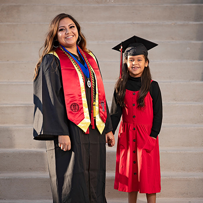 A woman in a black graduation gown stands and holds hands with her daughter who wears her mother's graduation cap.