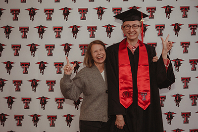A young man stands next to his mother wearing a graduation cap and stoll and while they both perform the guns up symbol.