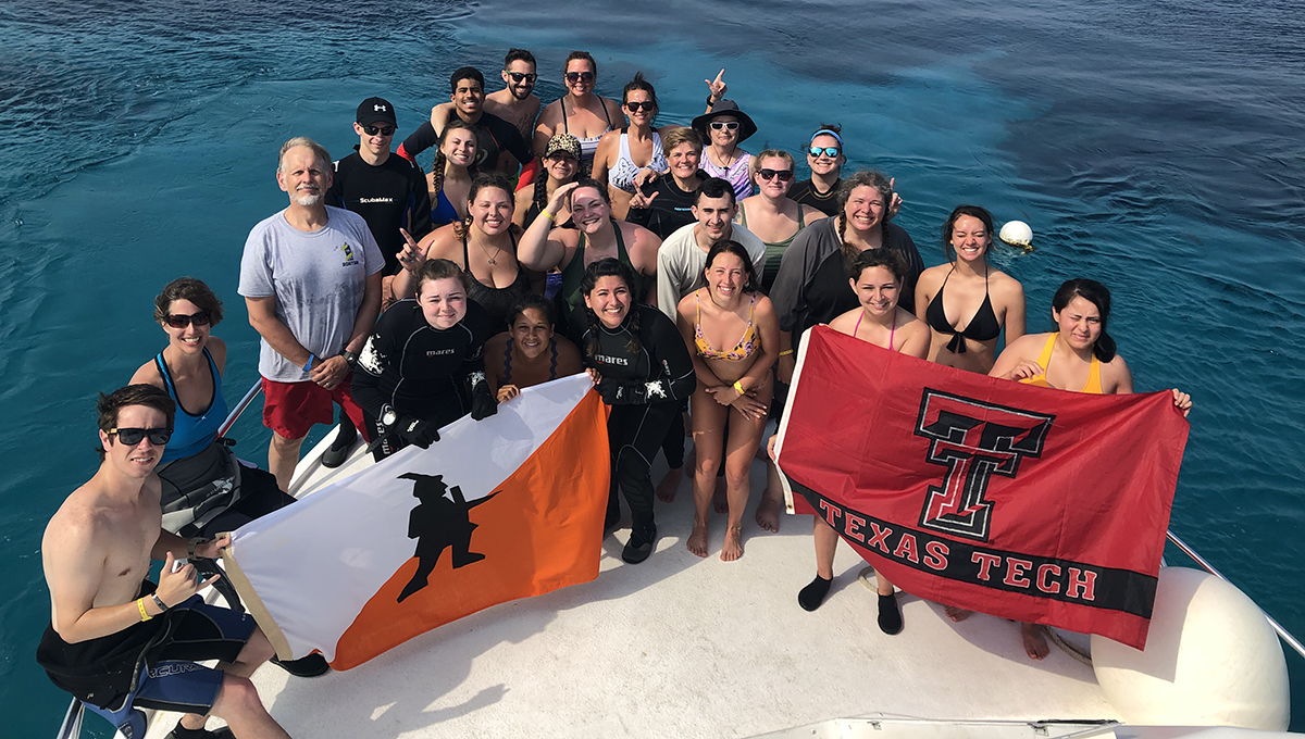 A group of men, women, and young students stand together on a boat that rest on top of clear blue water and look up at the camera while a few students hold both a texas tech flag and an unknown flag.