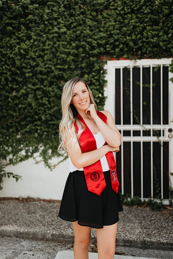 Sarah stands outdoors wearing a white shirt and black skirt with a ttu stole around her neck as she poses with her right arm across her body and her left arm and fist under her chin smiling at the camera