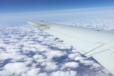 The left wing of an airplane as seen from a plane window as it hovers above white clouds and a blue sky