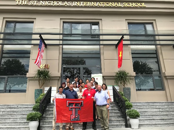 TTU K-12 staff stand alongside St, Nicholas personnel on the steps in front of the new St. Nicholas International School while holding a Texas Tech University flag.
