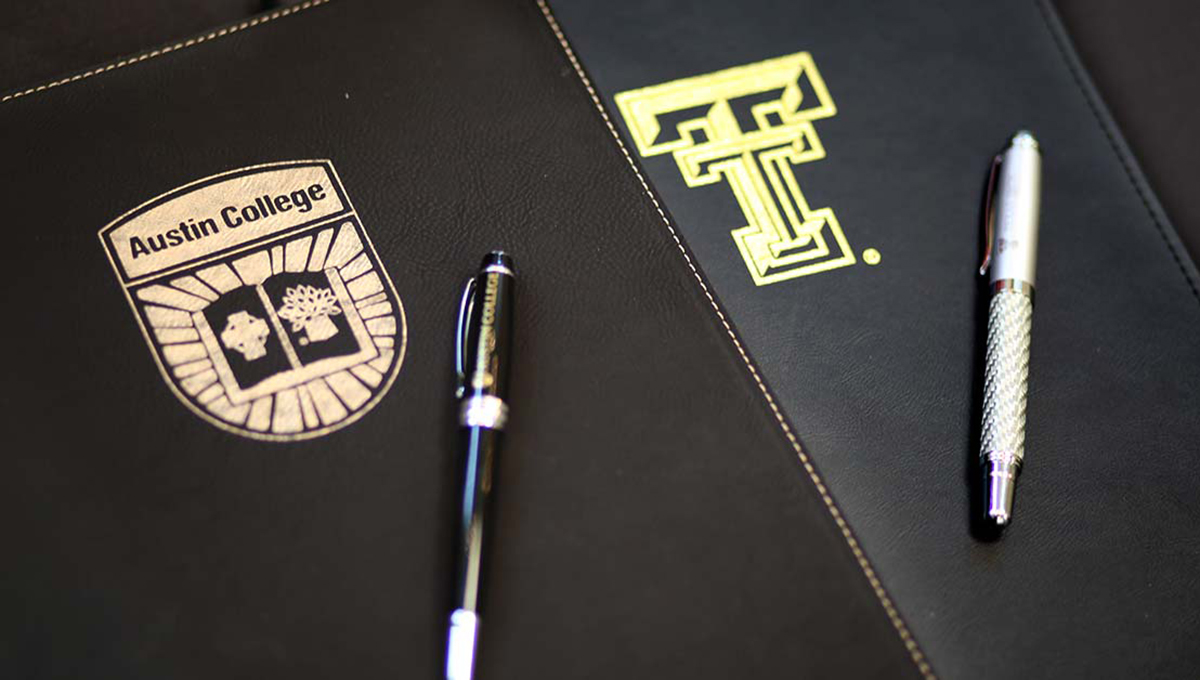 A black binder with the Austin College logo on the front rests on top of another black binder with the Texas Tech University logo and two writing pens lay on each binder