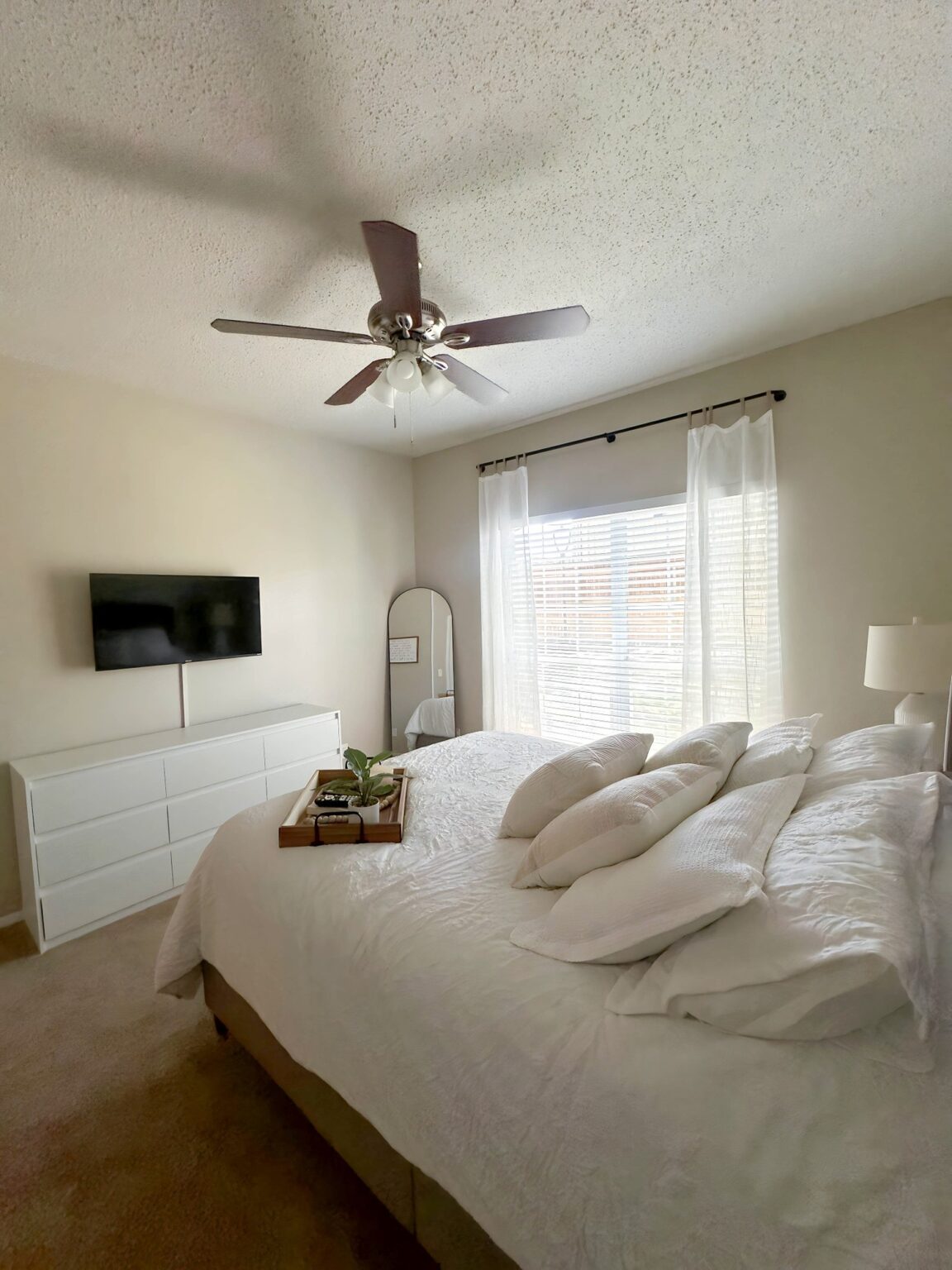 Bedroom with white bedding, dresser, wall-mounted television, ceiling fan, and large window.