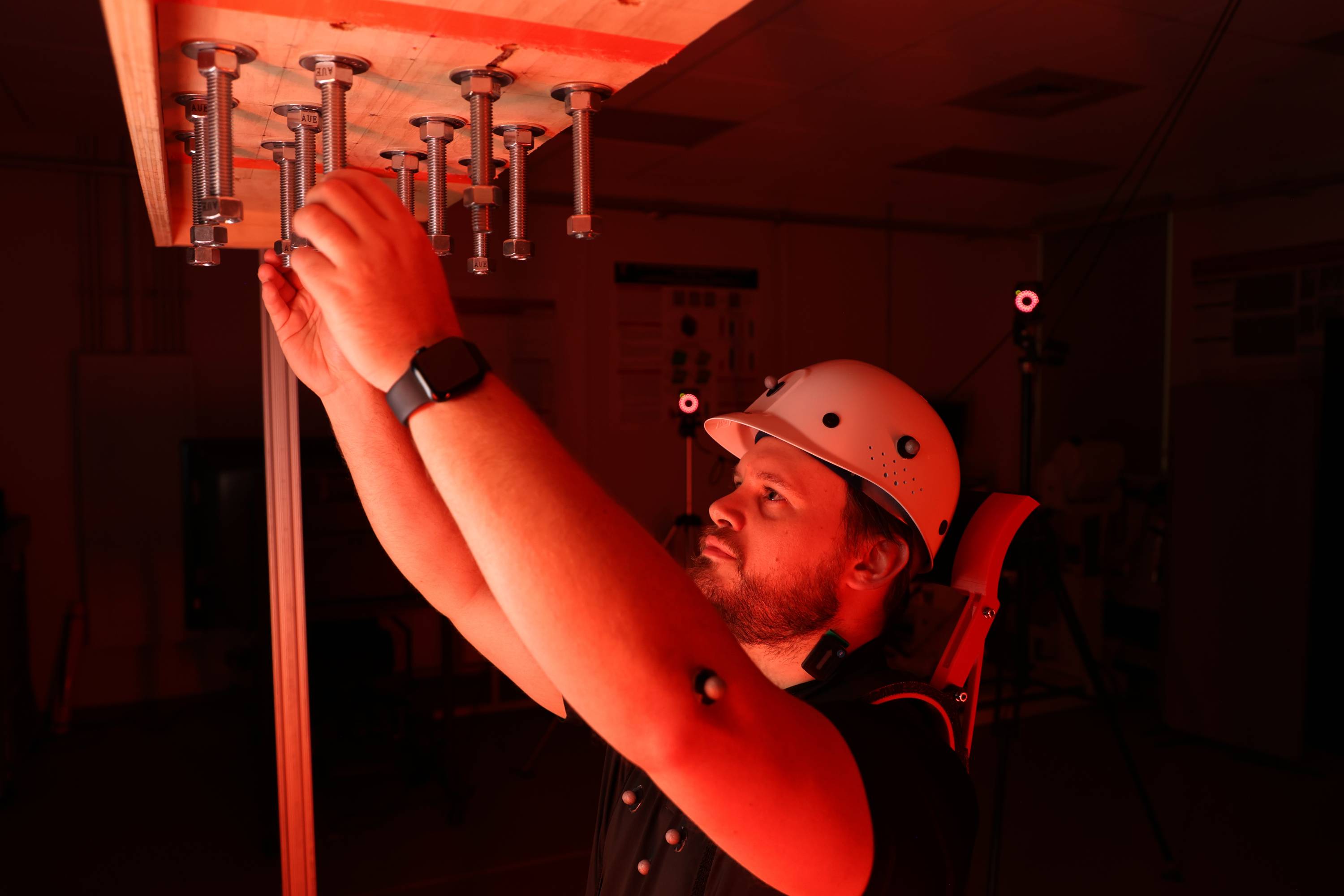 Student in research lab wearing a hard hat, reaching up to screw in bolts in a wooden plank above the head. The student is wearing a neckbrace in order to research neck strain when reaching above the head at work.