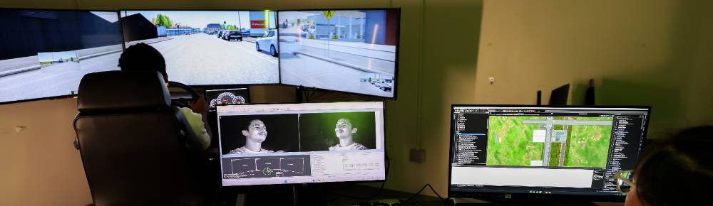 student sitting at driving simulator with three large screens. Behind the student is two computer monitors looking at their facial and eye movement and the driving map to analyze human factors and interaction while driving.