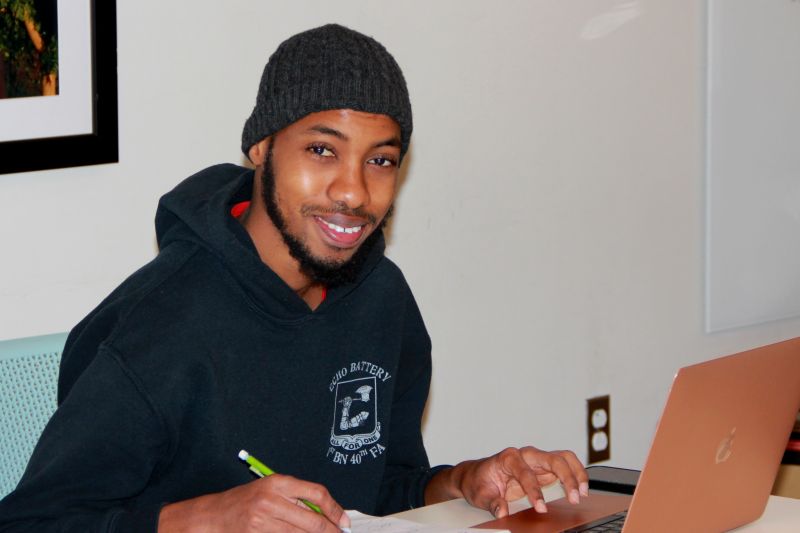 A student holding a pencil and writing on paper while smiling at the camera.