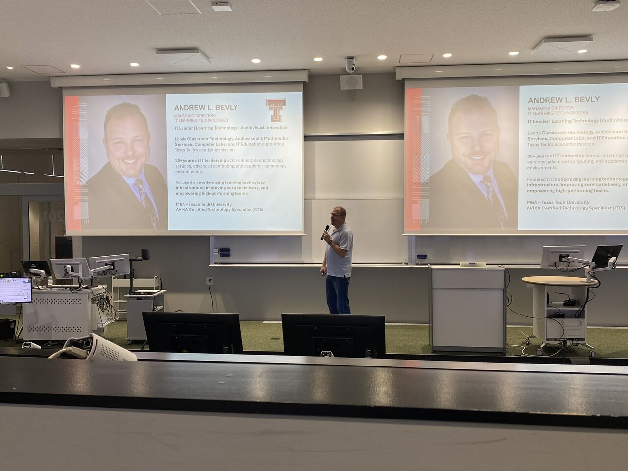Andrew speaking in a modern lecture hall with two large screens displaying a professional profile slide.
