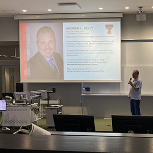 Andrew speaking in a modern lecture hall with two large screens displaying a professional profile slide.