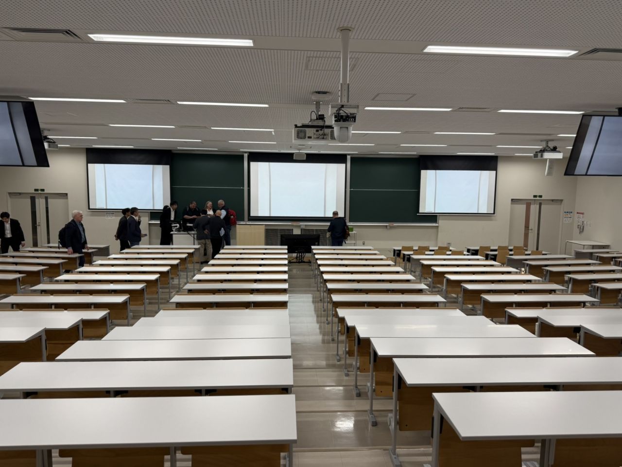 Spacious university lecture hall with rows of white desks and wooden chairs facing multiple large projection screens and chalkboards.