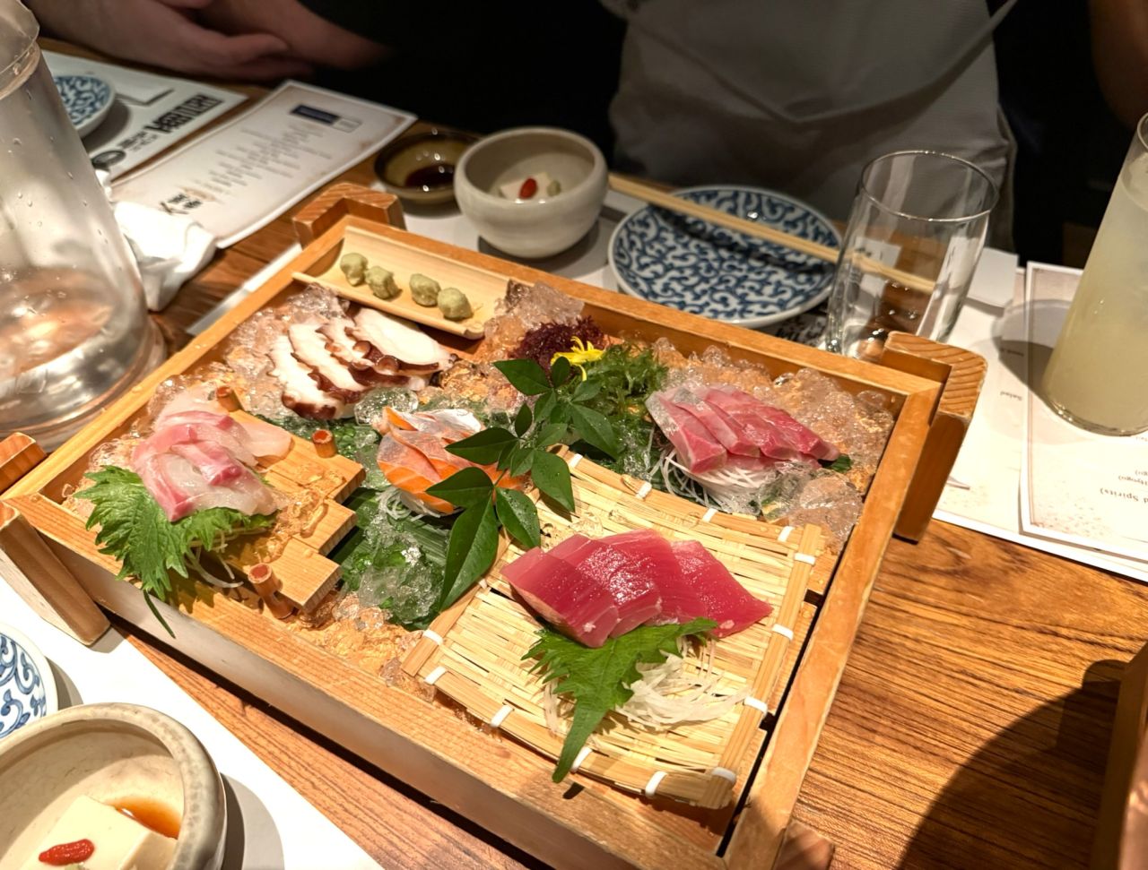 Assorted sashimi platter on crushed ice with fresh fish slices, garnished with green leaves and wasabi, served on a wooden tray.