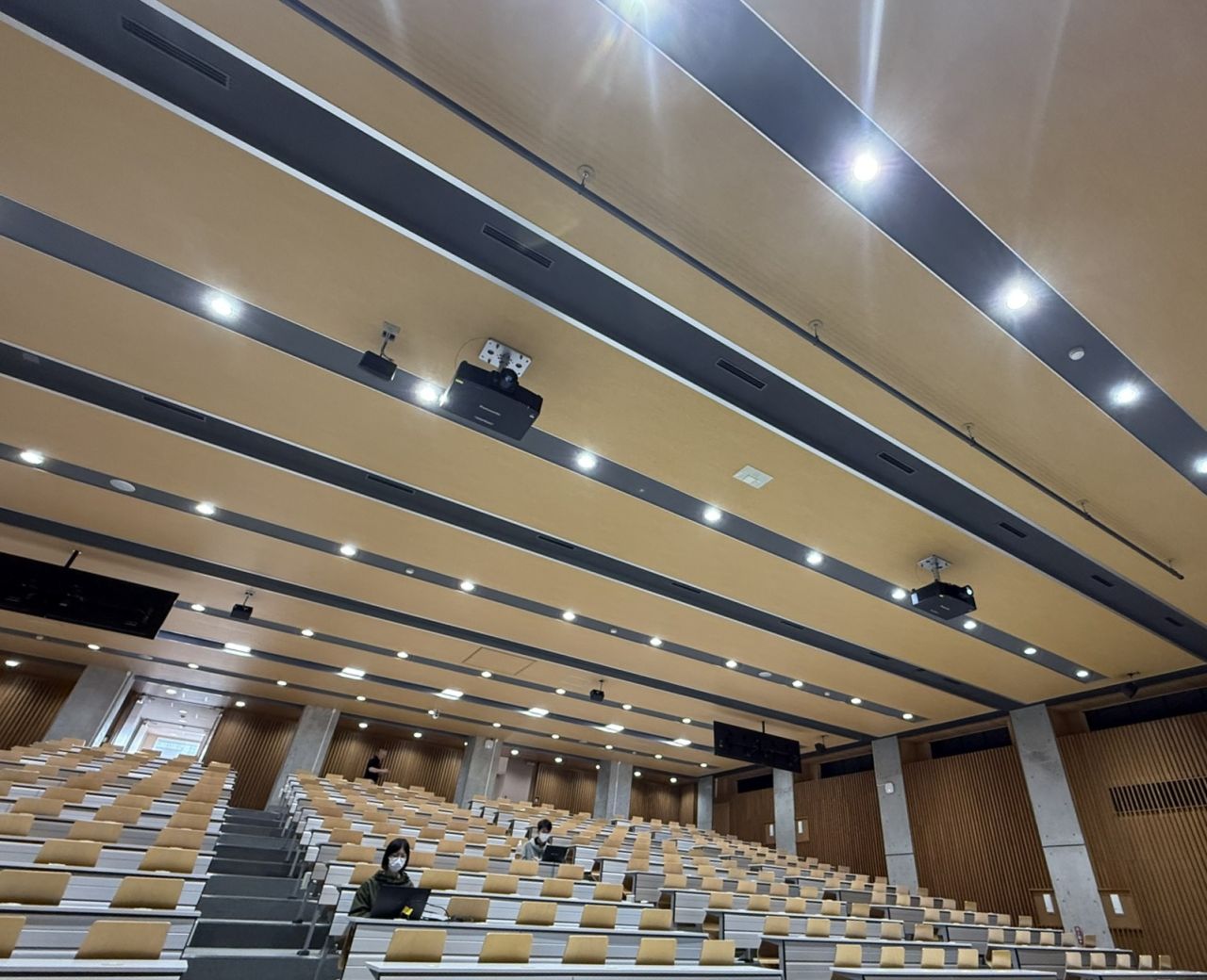  Large modern lecture hall with tiered seating and a few students spaced out wearing masks.