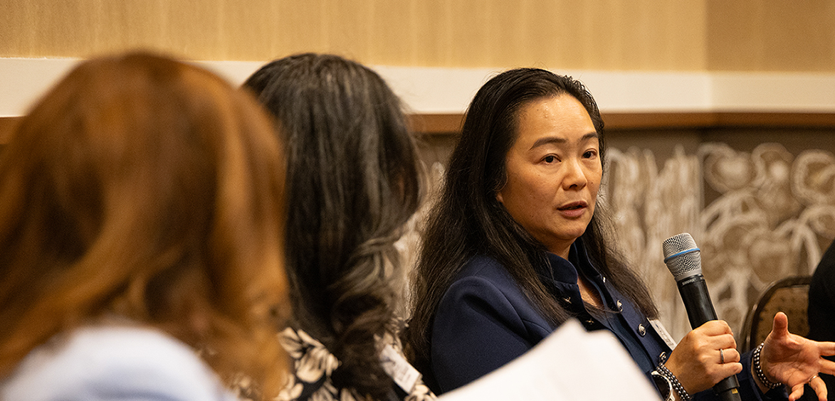 Yung Ng speaking into a microphone as a panelist at the West Texas Women in Technology Luncheon, gesturing while seated alongside fellow panelists.