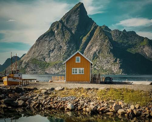 A small house sits next to a boat dock along a rocky shoreline. A large mountain towers above the house in the background.